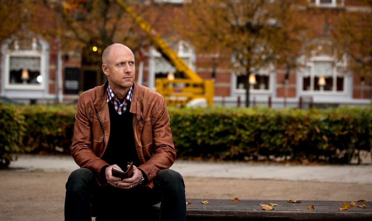 A thoughtful man in a leather jacket sits on a bench in an urban setting during fall.