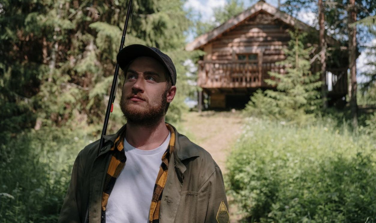 Bearded man holding fishing rod in lush greenery by a log cabin.