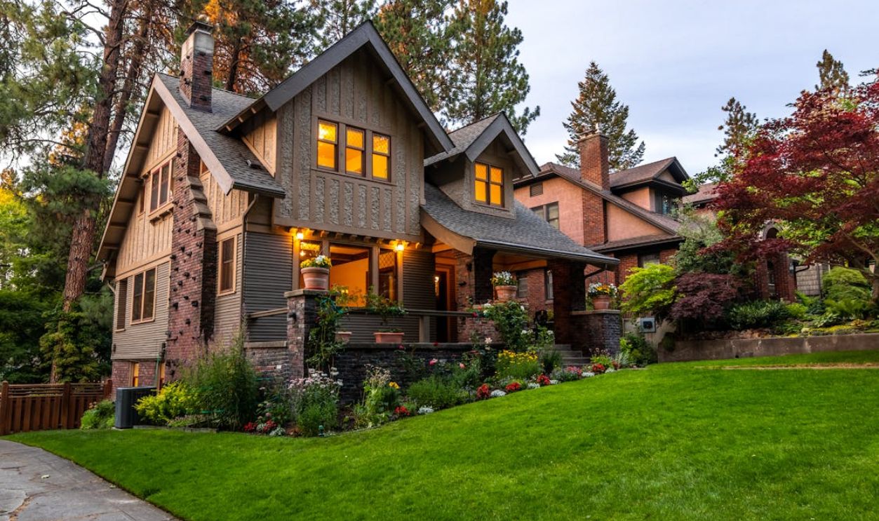 A beautifully lit Victorian-style house surrounded by a lush garden at twilight, showcasing classic architecture.