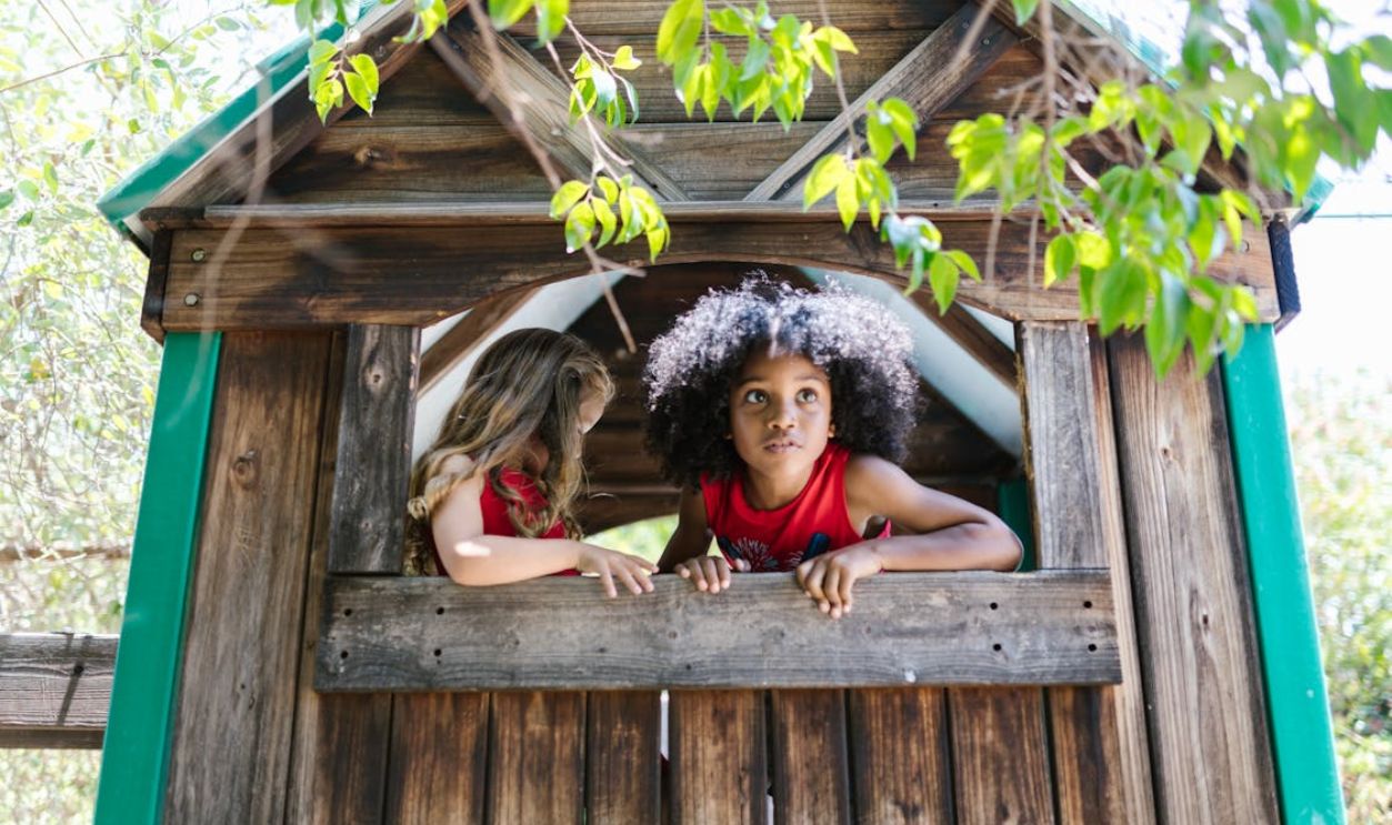 Two children exploring and playing in a wooden treehouse surrounded by green leaves.
