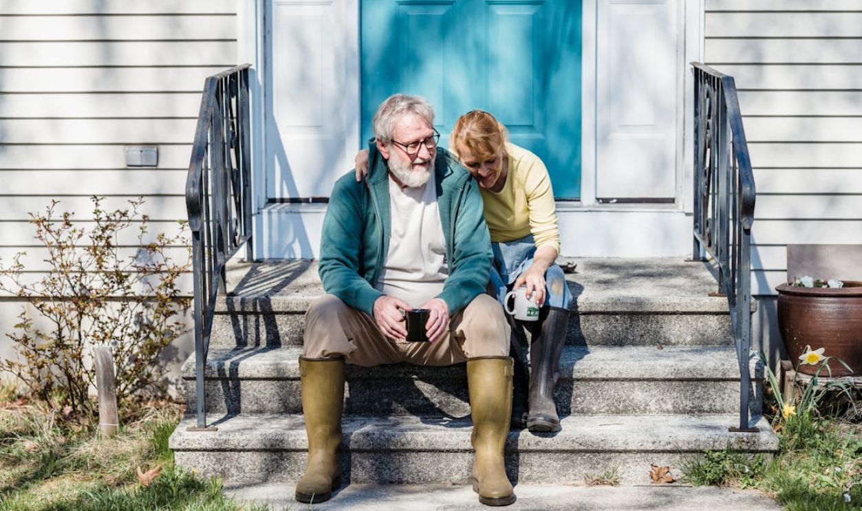 An elderly couple sitting affectionately on their porch steps, sipping coffee and enjoying a sunny day.