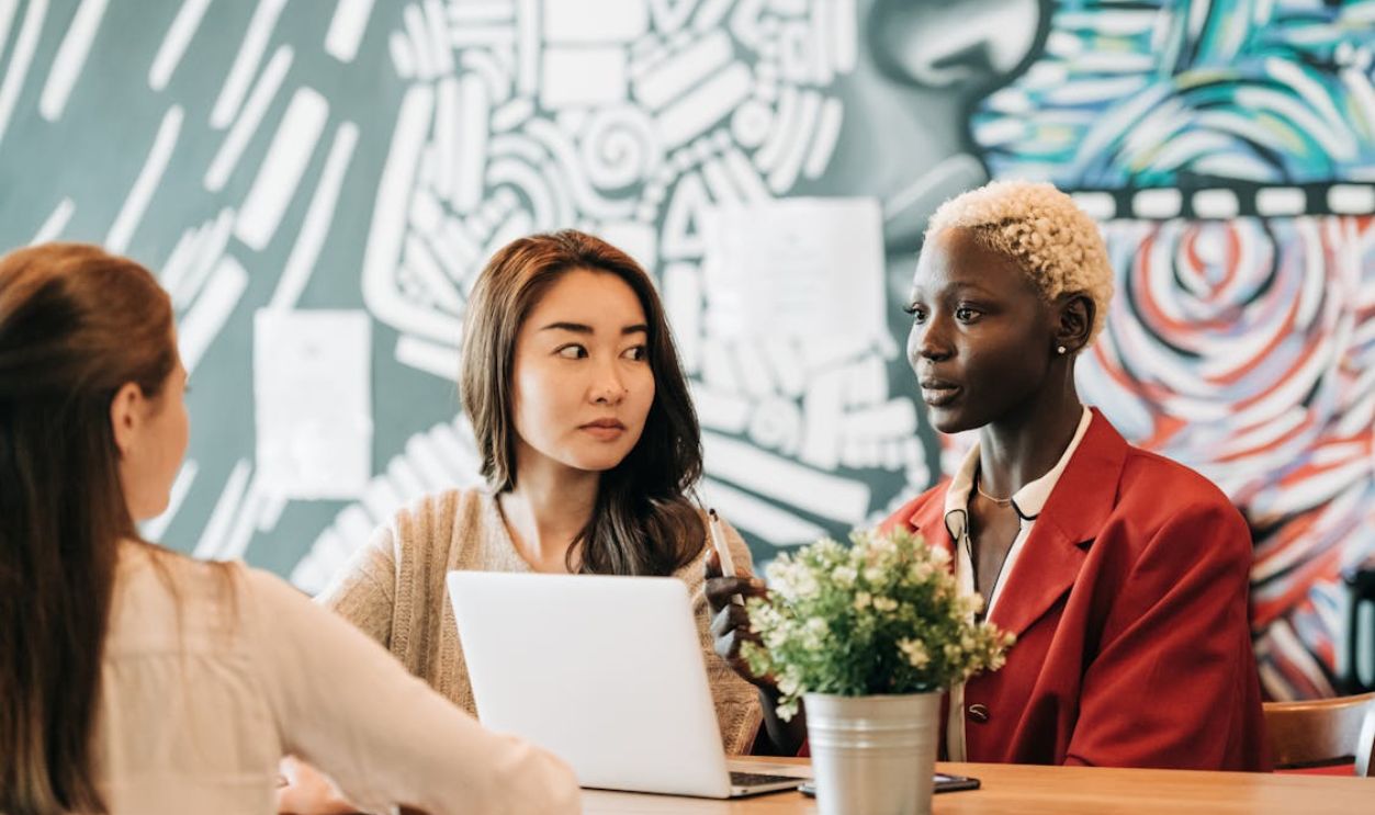 Three women collaborating using a laptop in a trendy cafe with vibrant mural.