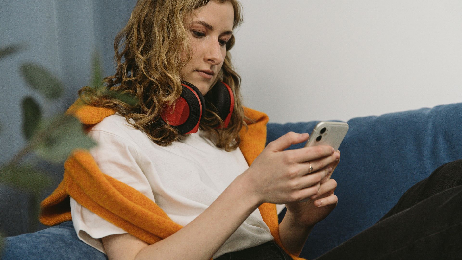 Young woman sitting on a couch using her smartphone, wearing headphones, with a relaxed expression.