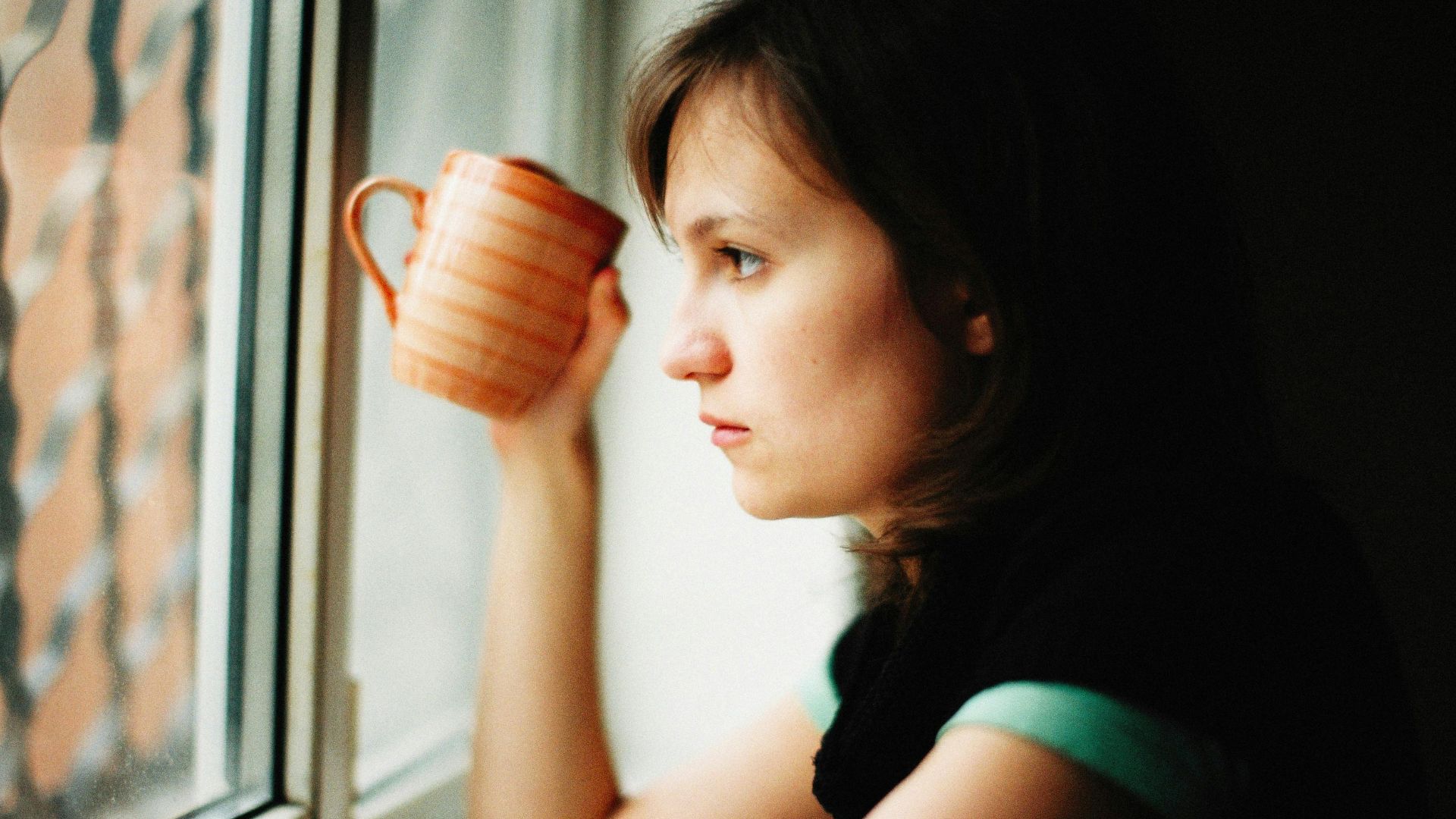 A woman holding a mug gazes thoughtfully out the window, adding mood to the scene.