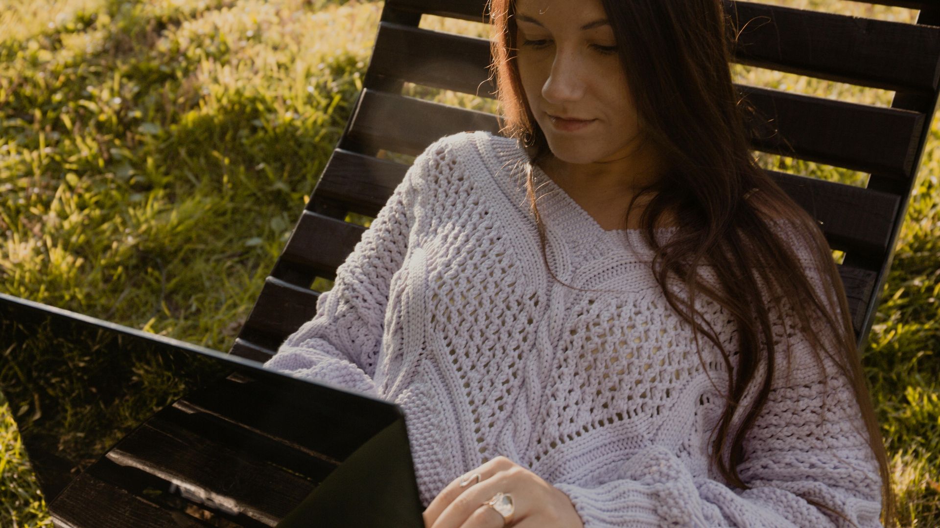 Woman in knitwear using laptop outside on a sunny day, enjoying leisure time.