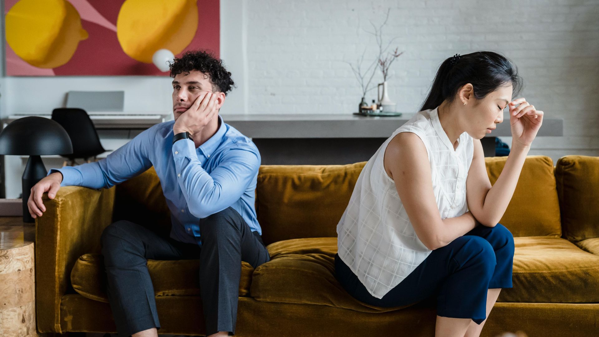 A couple sits apart on a sofa, looking unhappy and deep in thought, reflecting relationship conflict.
