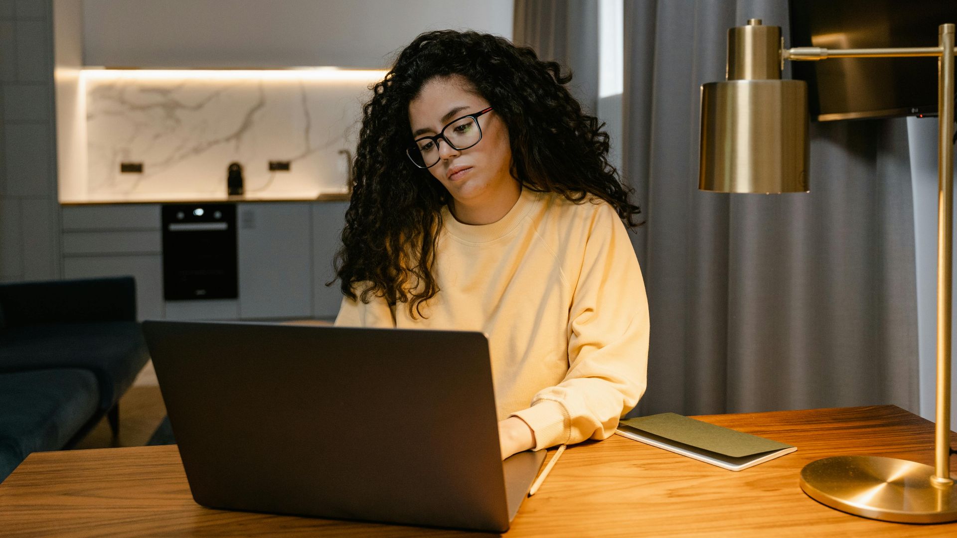 Woman in a yellow sweatshirt using a laptop at home, focusing on remote work in a cozy, modern interior.