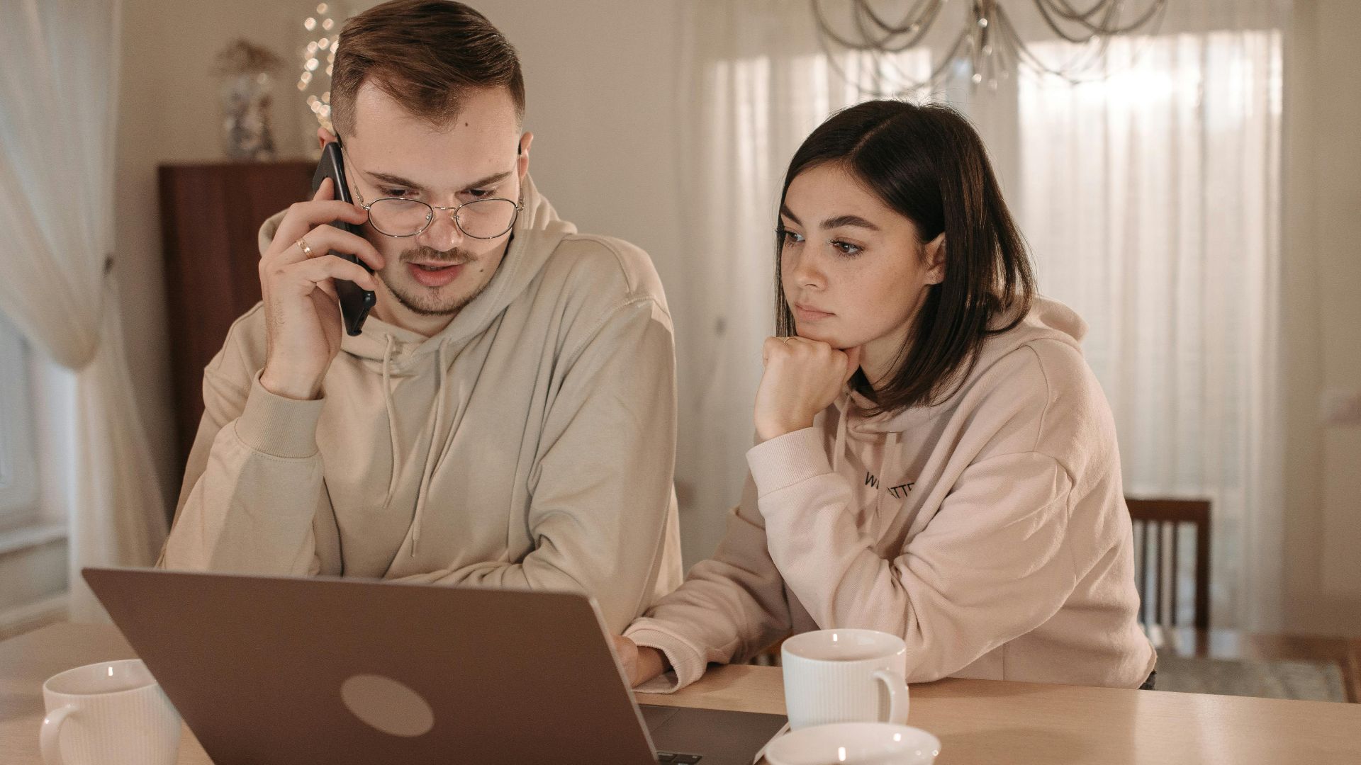A couple in hoodies focused on work using a laptop and smartphone at home.
