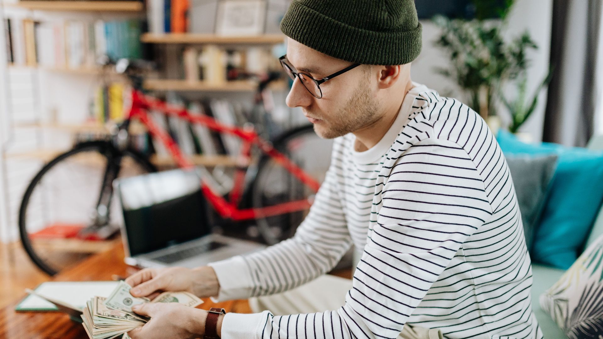 Man in striped shirt counting cash indoors, with a bicycle in the background.