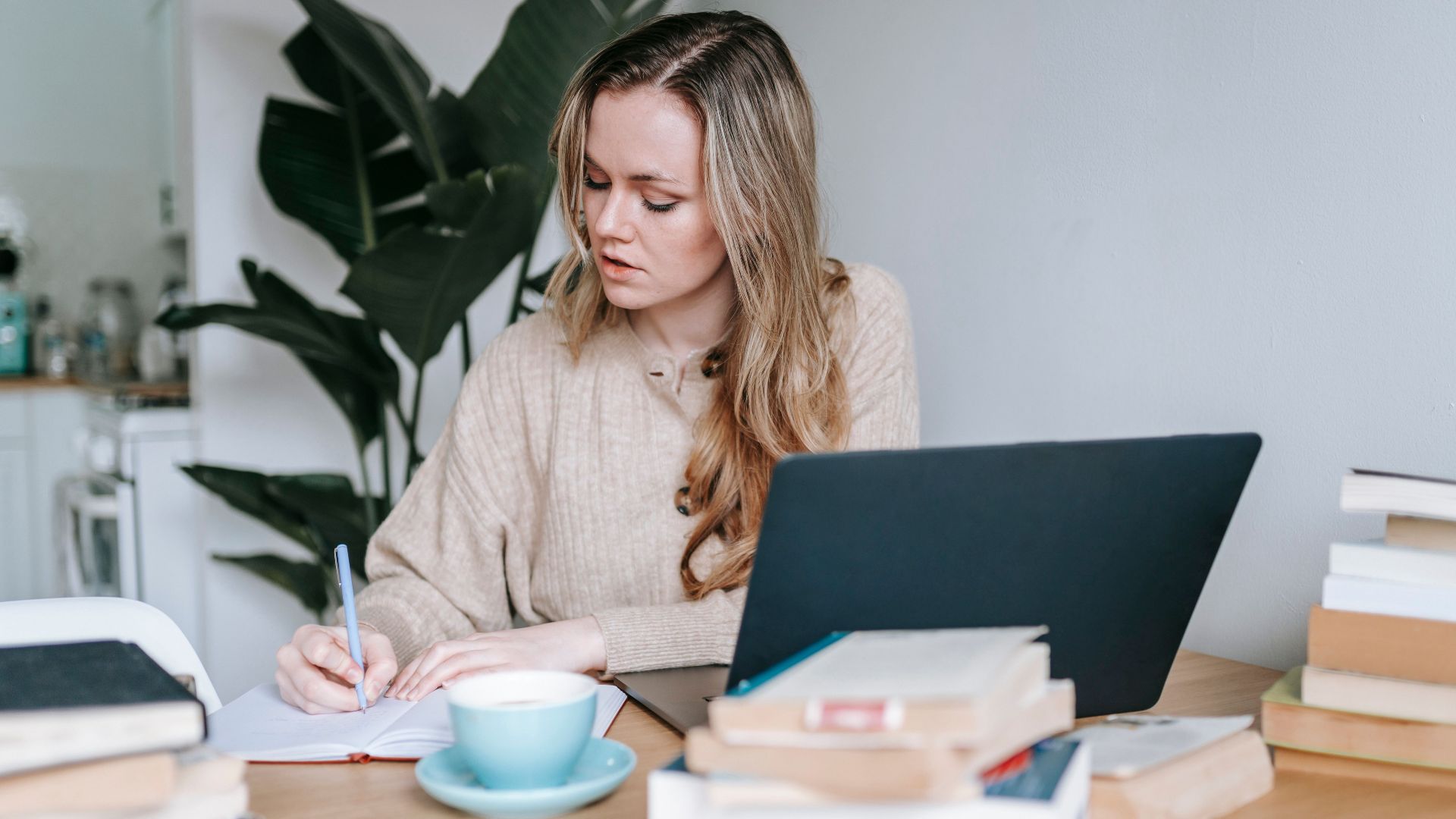 Young focused female with long hair taking notes at table with netbook and pile of books