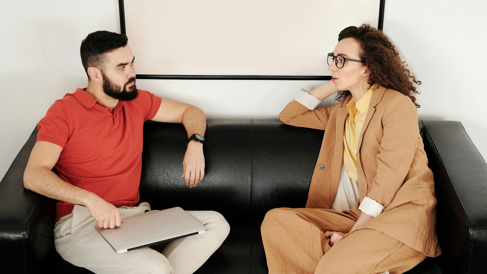 A man and woman sit on a black sofa, having a professional conversation.