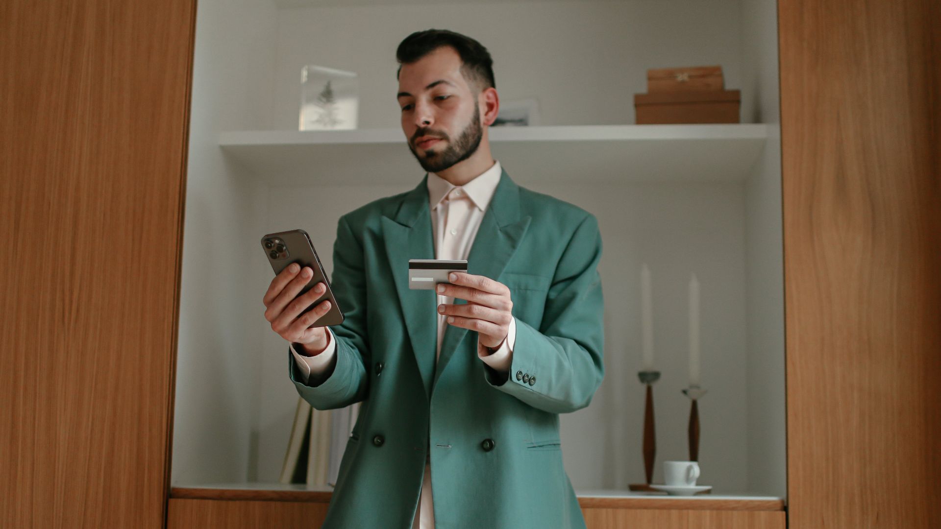 A bearded man in a green suit is using a smartphone and credit card indoors, showcasing modern technology and lifestyle.