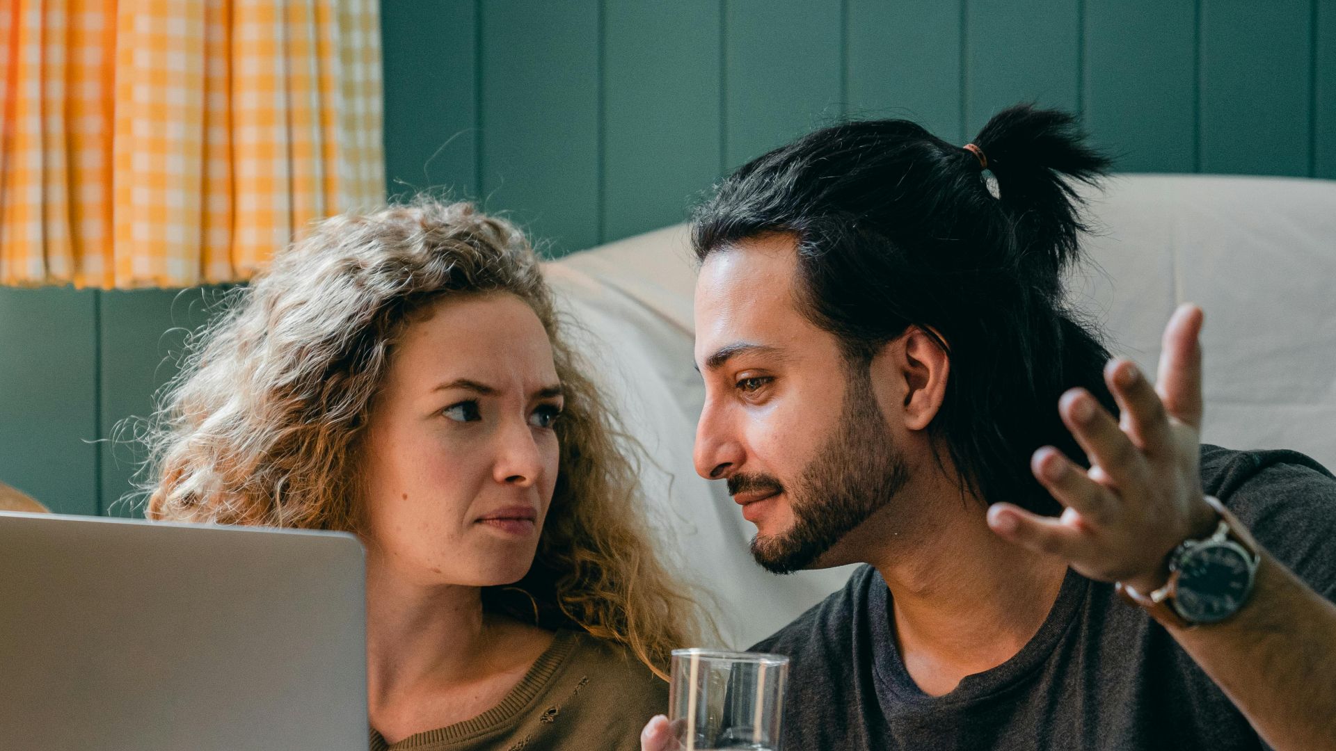 Bewildered young multiethnic couple in casual outfits sitting on floor against blue wall while browsing modern netbook and discussing news