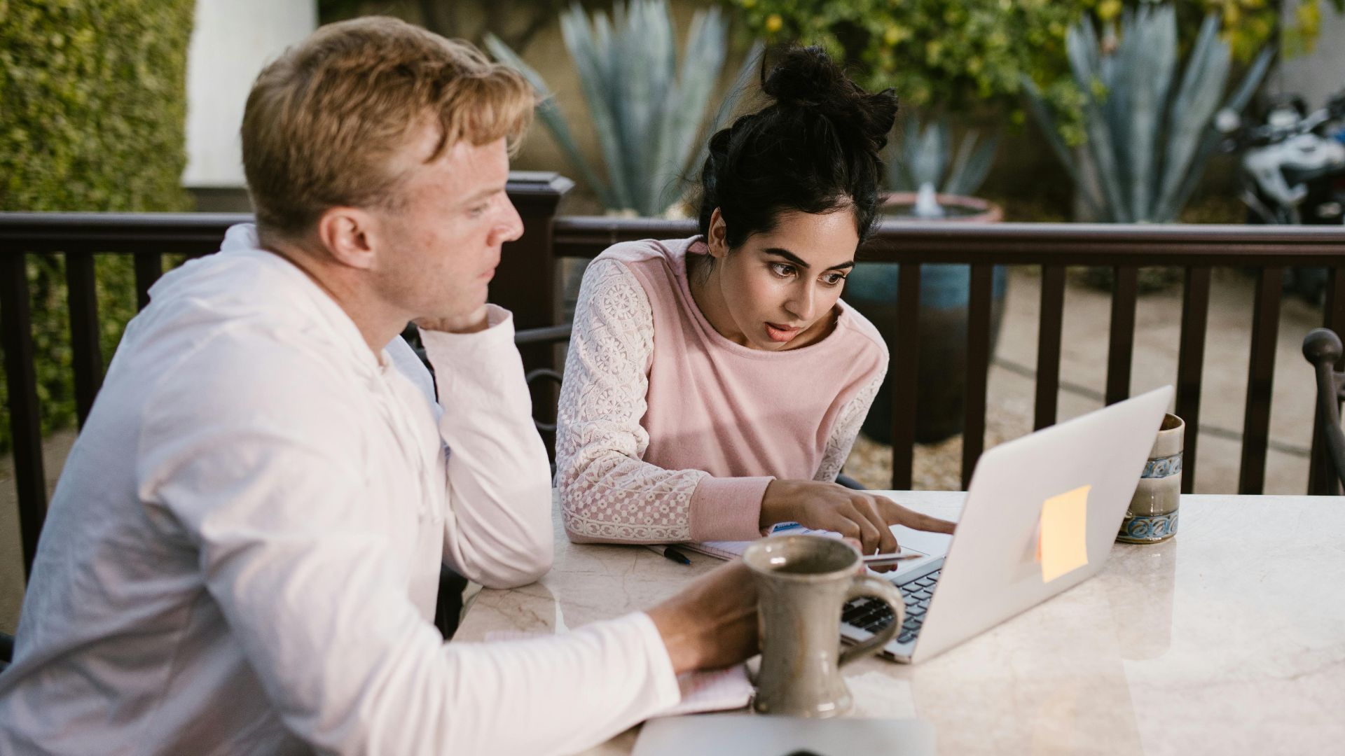 Two adults collaborating on a laptop, seated outdoors with coffee on the table.