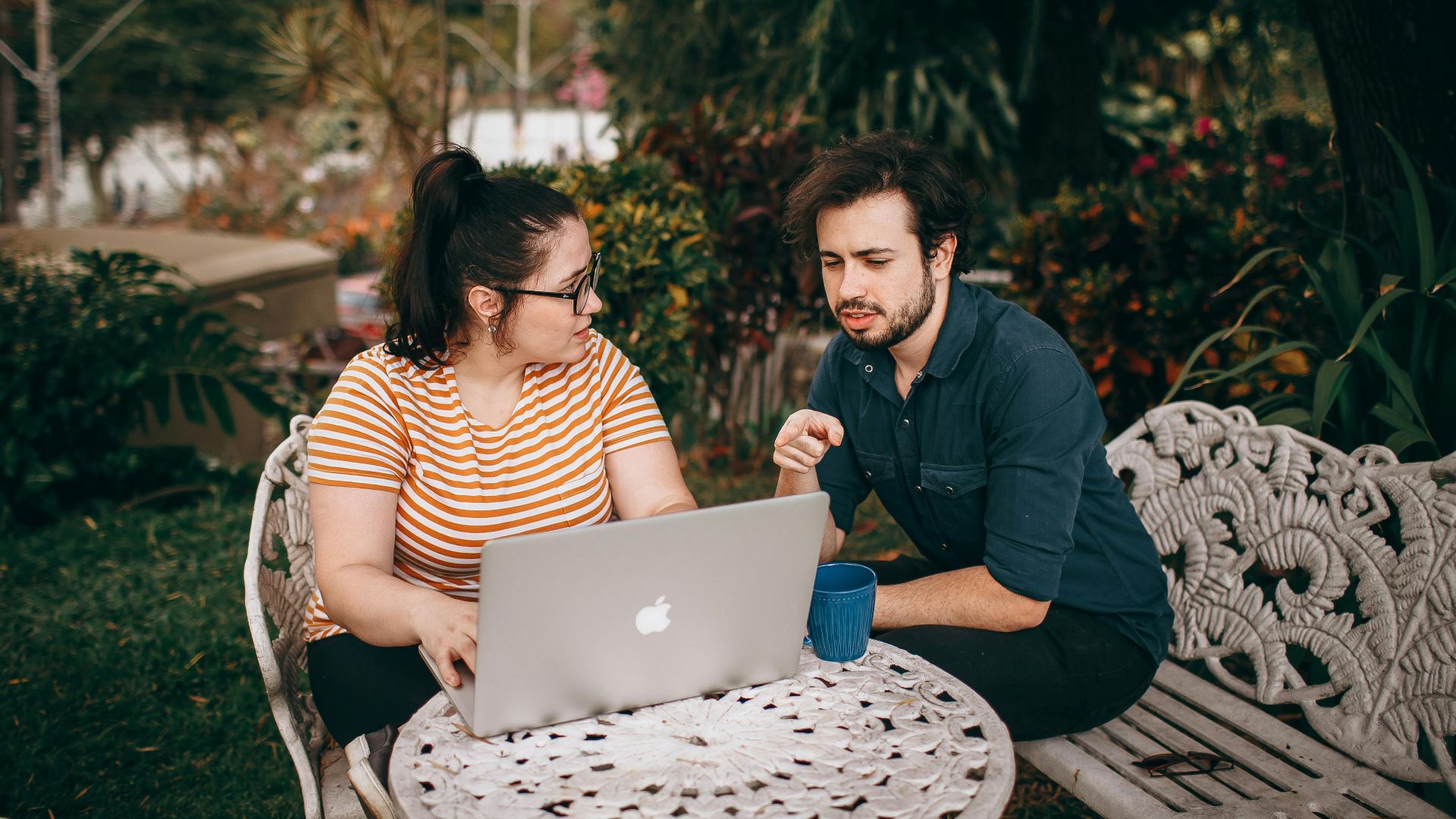 A man and woman working together on a laptop outdoors in a lush garden.