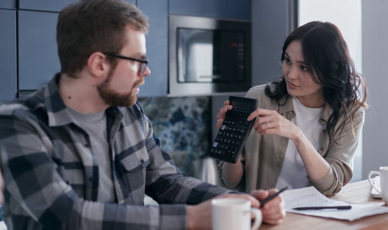 A couple discussing finances with a calculator and papers at a wooden table, looking concerned