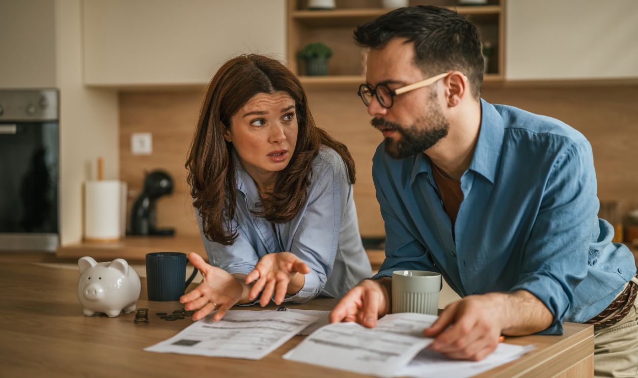 Couple at kitchen counter stressed over household bills and debt, surrounded by paperwork, calculator and piggy bank as they discuss budgeting, payments and finances