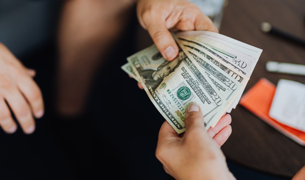 A close-up view of hands exchanging dollar bills indoors, symbolizing payment