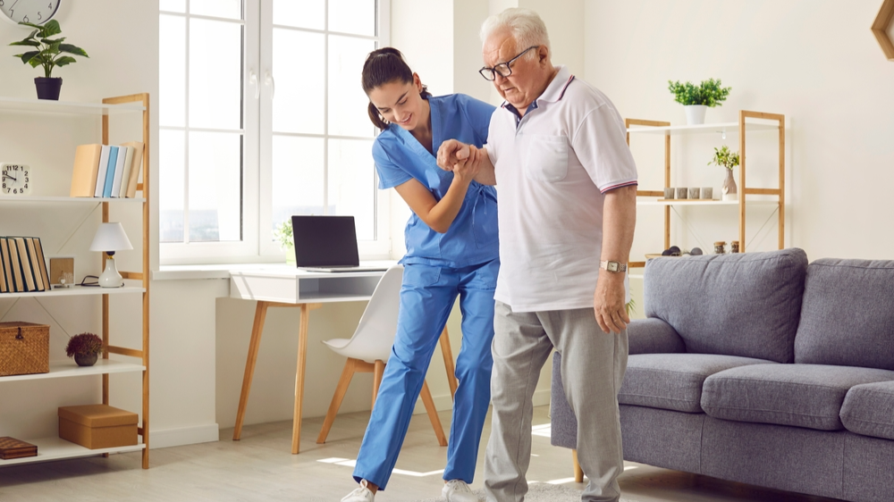 Young nurse helping elderly man walk in the room, holding his hand, supporting him. Treatment and rehabilitation after injury or stroke, life in assisted living facility, senior care concept