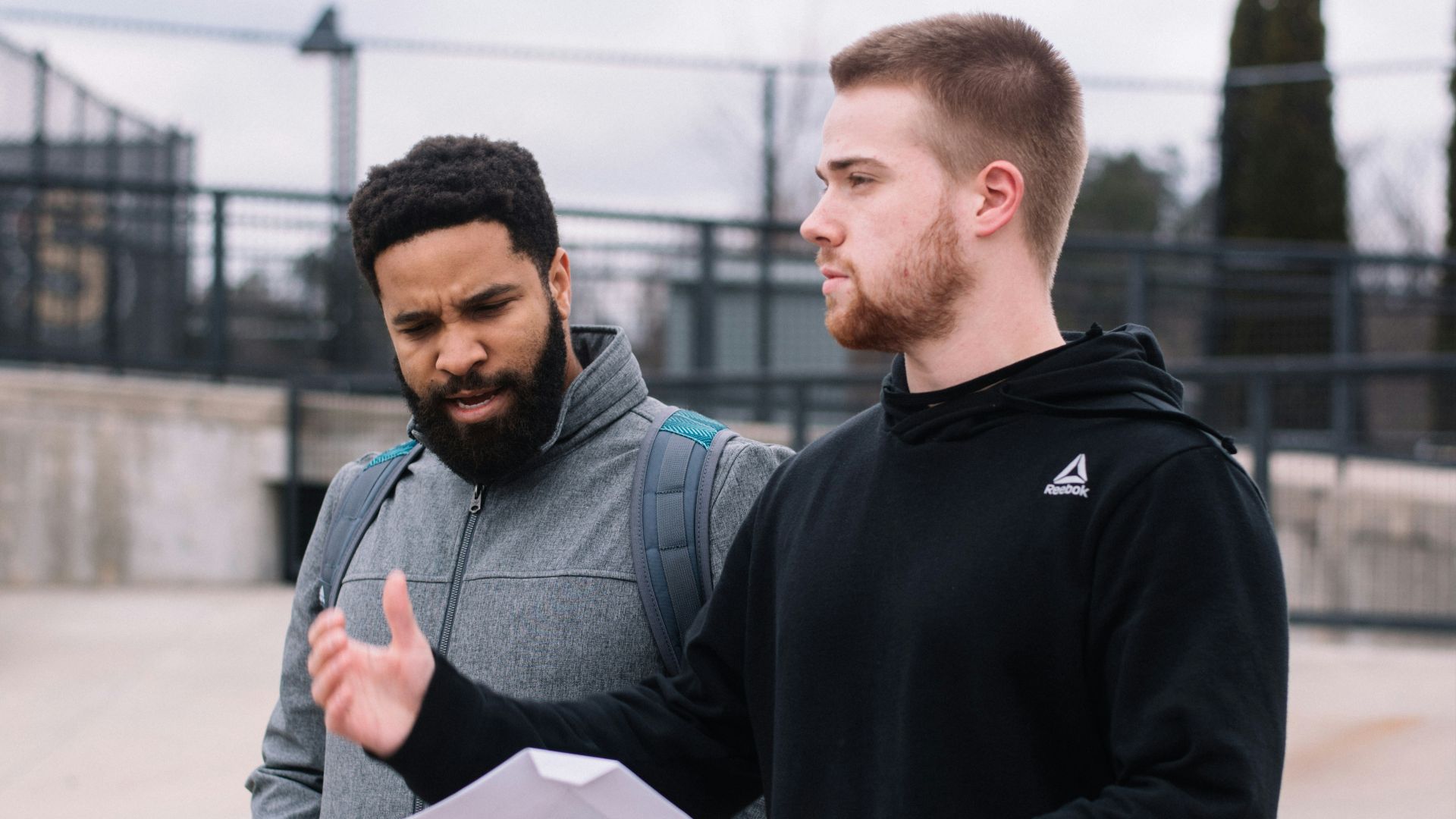 man in black hoodie holding white printer paper and man in grey zip-up jacket during daytime