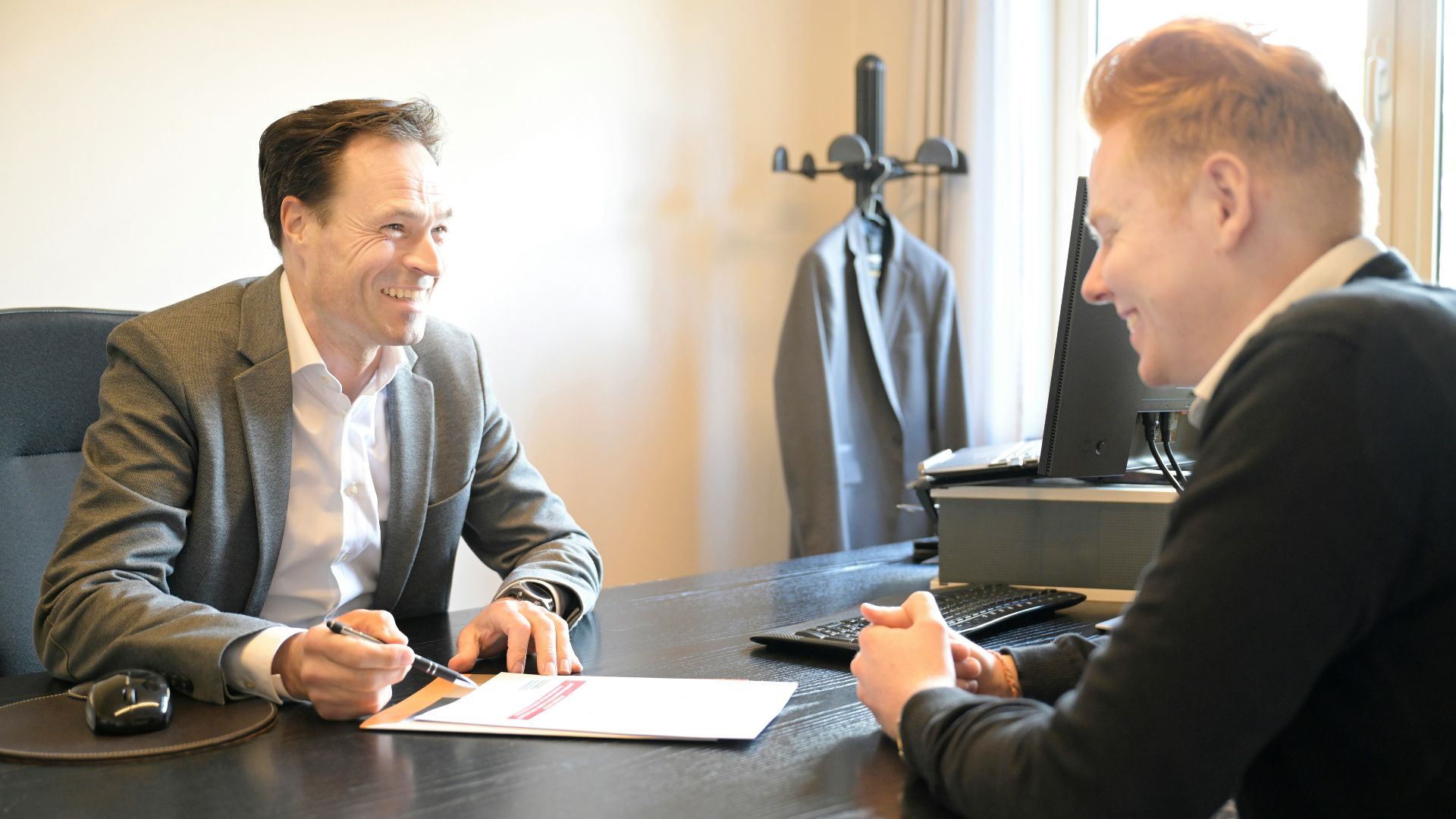 Two smiling men discussing documents at an office desk.