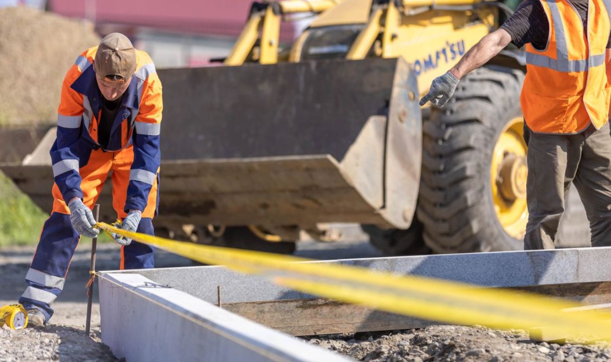Construction workers using measuring tape at a sunny outdoor site with a bulldozer in the background.