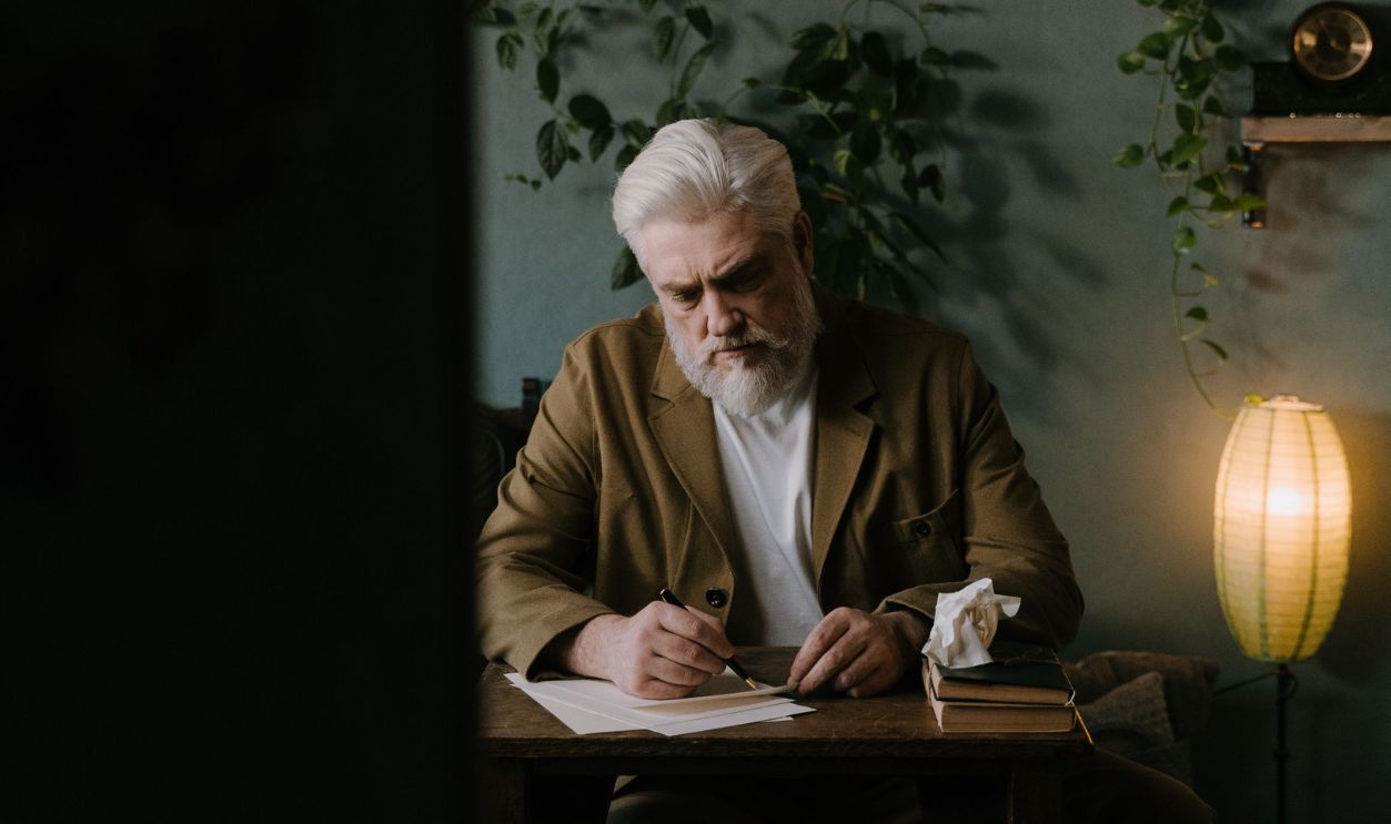 Elderly man with white hair and beard writing at a wooden desk in a warmly lit room with plants
