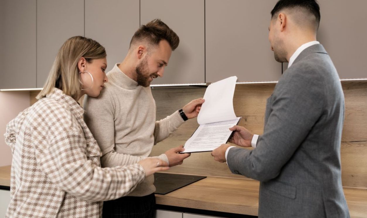 A couple reviews real estate documents with an agent in a modern indoor setting, discussing a potential property purchase.