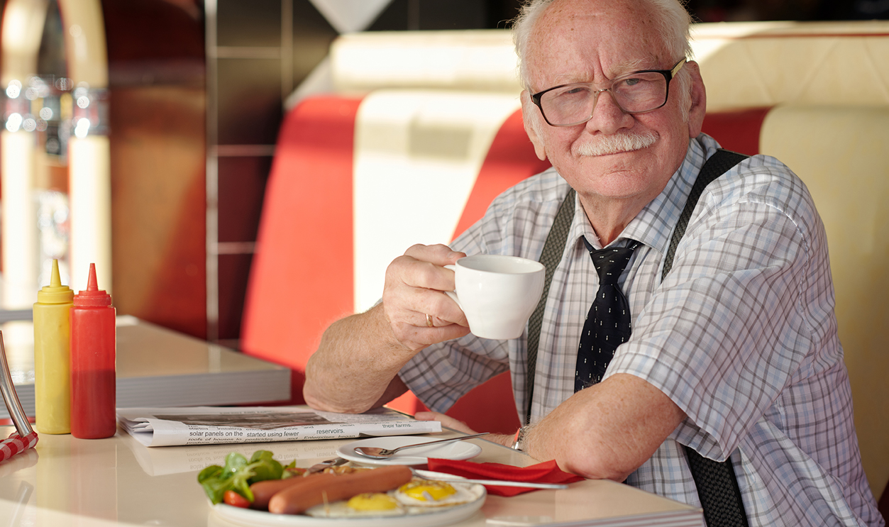Portrait of elderly man with mustache sitting in diner, smiling while holding coffee cup, with food on table and red, white booth in background