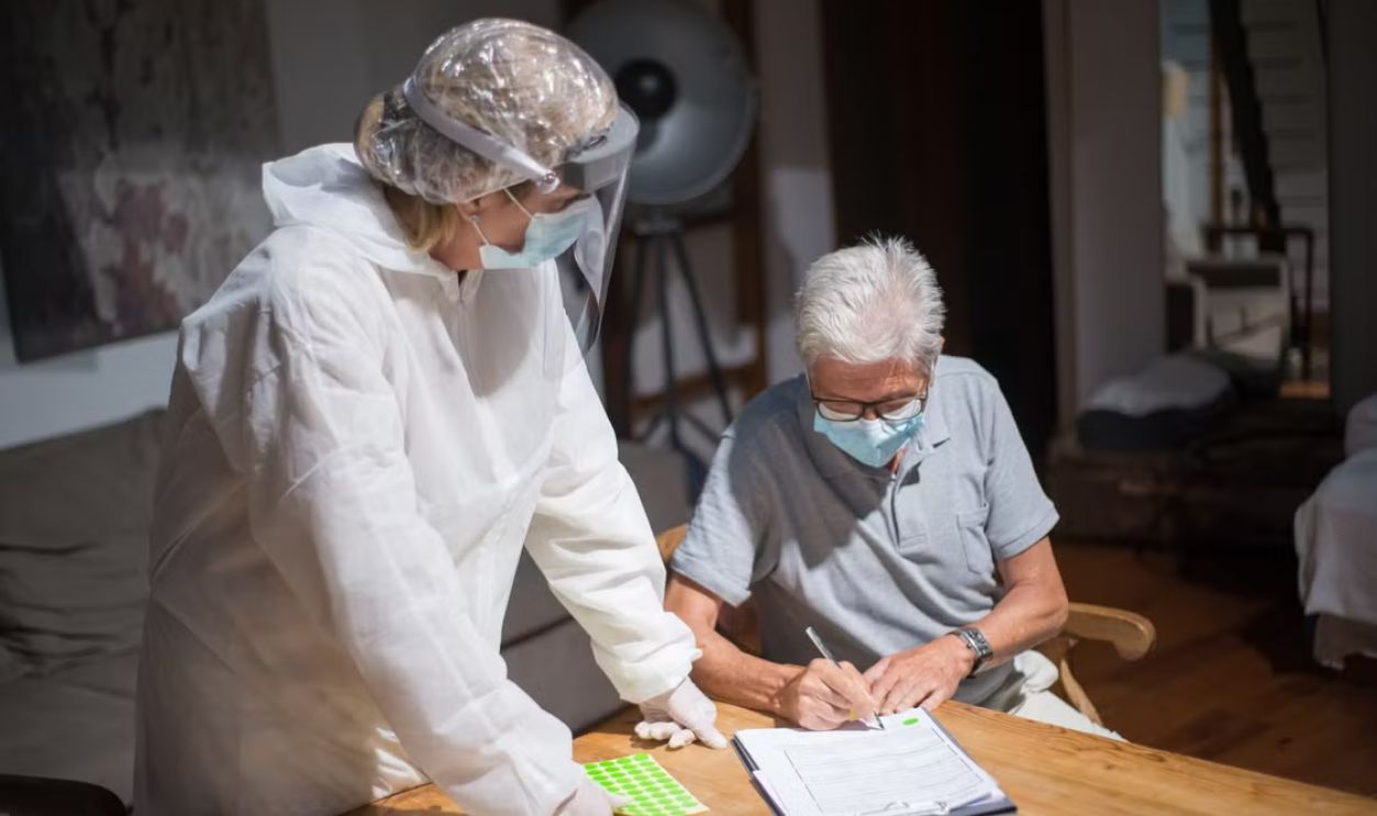A healthcare worker in protective gear assists an elderly man with paperwork in a home setting.