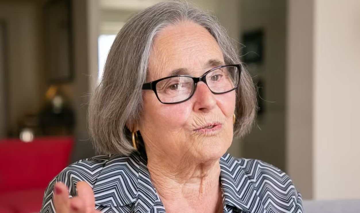 A senior caucasian woman with glasses speaking animatedly indoors.