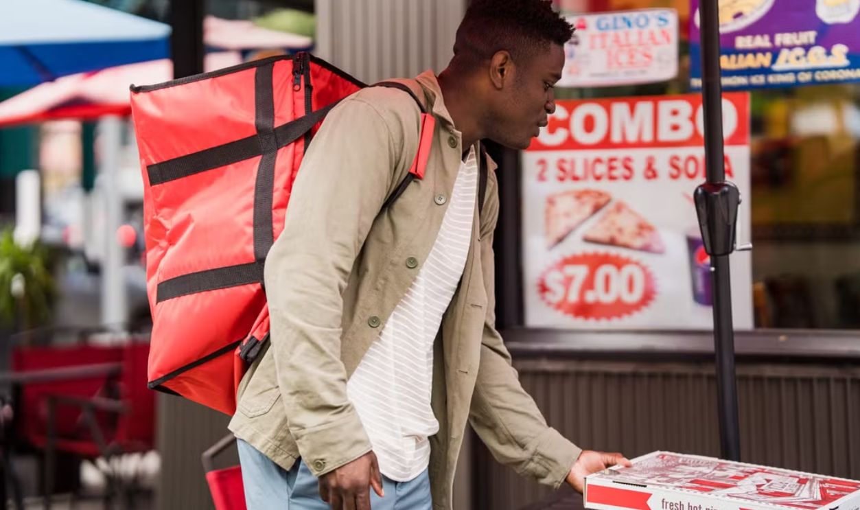 A pizza delivery person with a red backpack collects an order from a pizzeria.