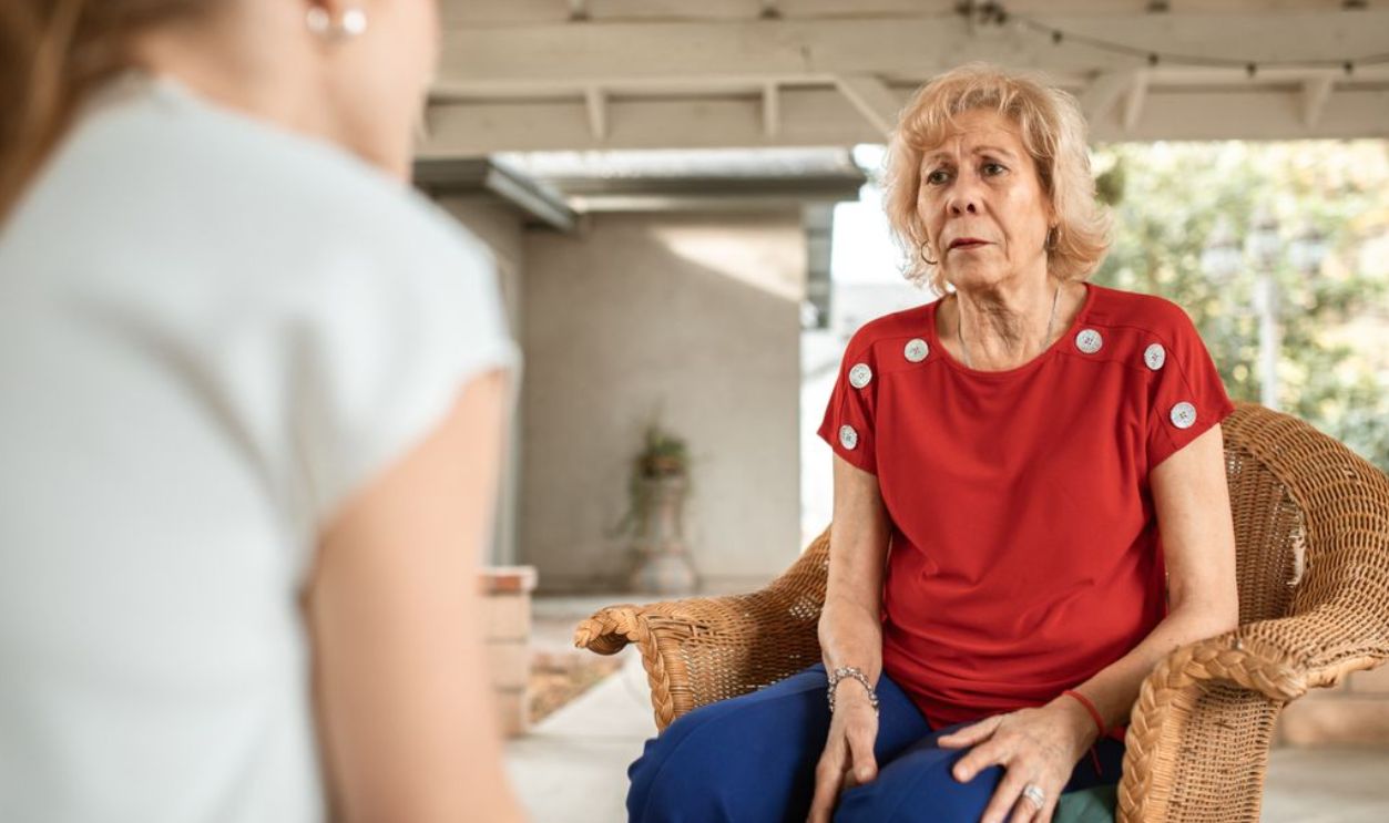 Senior woman in red shirt conversing indoors with younger woman. Daytime scene