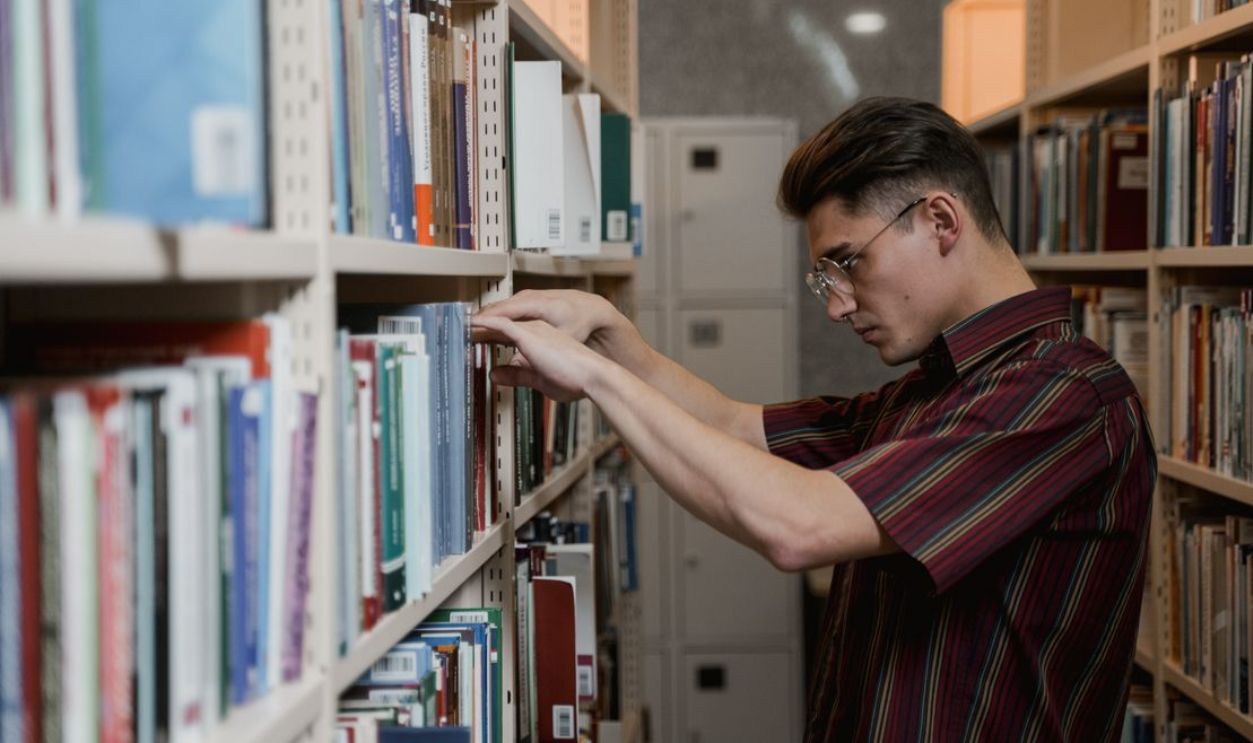 A young man in glasses searches library shelves for academic resources.