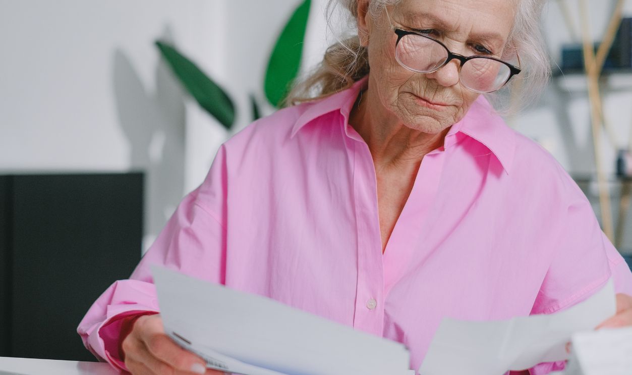 Elderly woman with eyeglasses reading papers at a desk, focusing indoors.