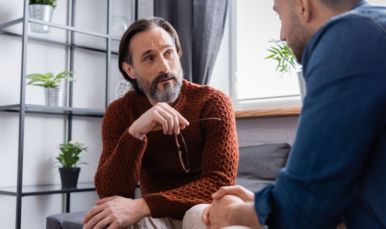 bearded man holding eyeglasses while listening to hispanic son on blurred foreground