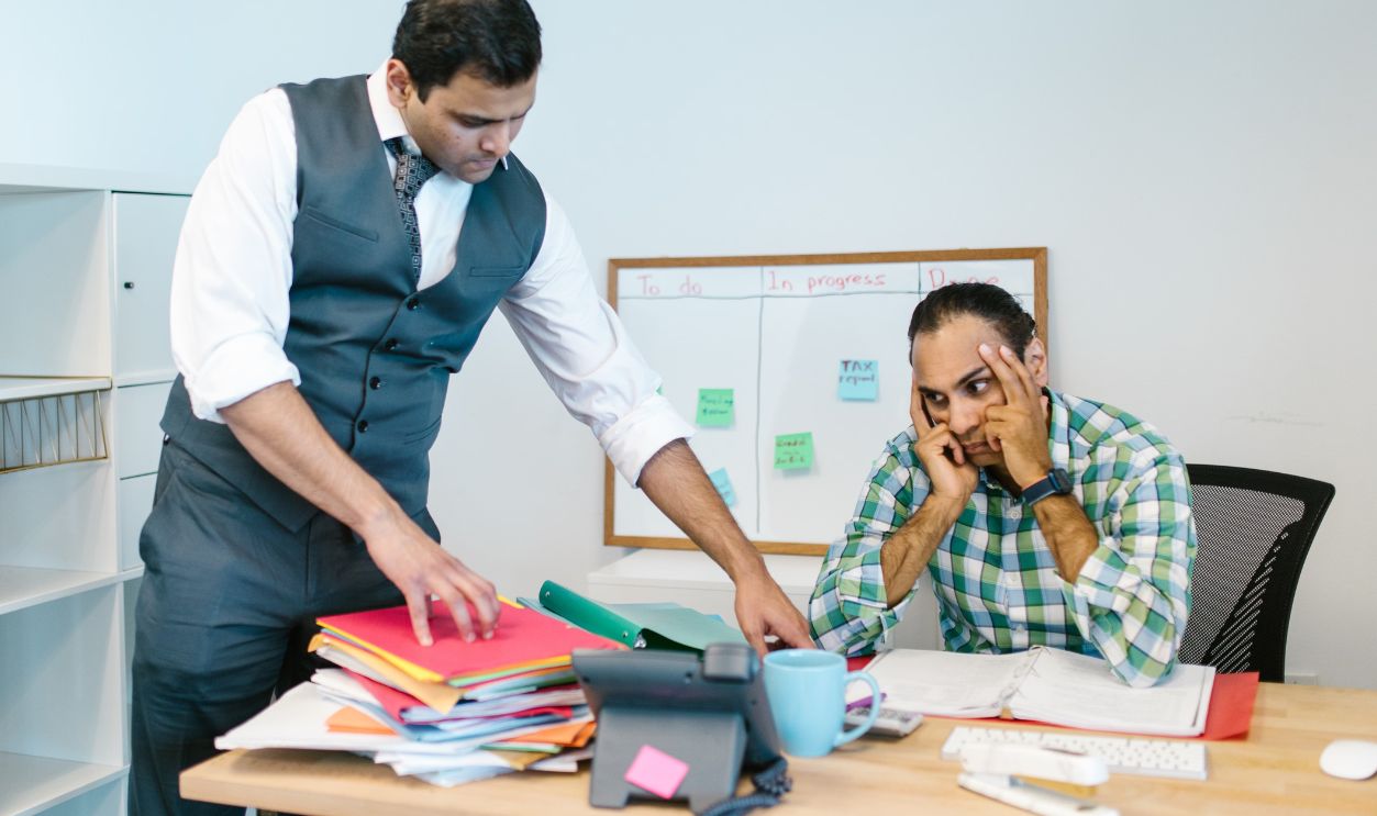 Two businessmen in an office, one stressed at the desk while another points at folders, discussing workload