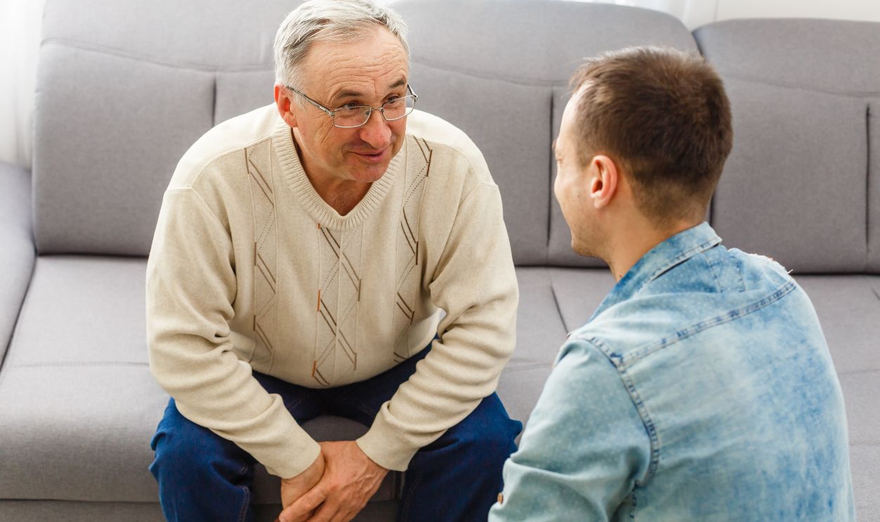 Smiling grown son sit on couch relax with senior dad talk sharing thoughts looking in eyes, happy millennial man rest on sofa speak with elderly father enjoy leisure family weekend at home.