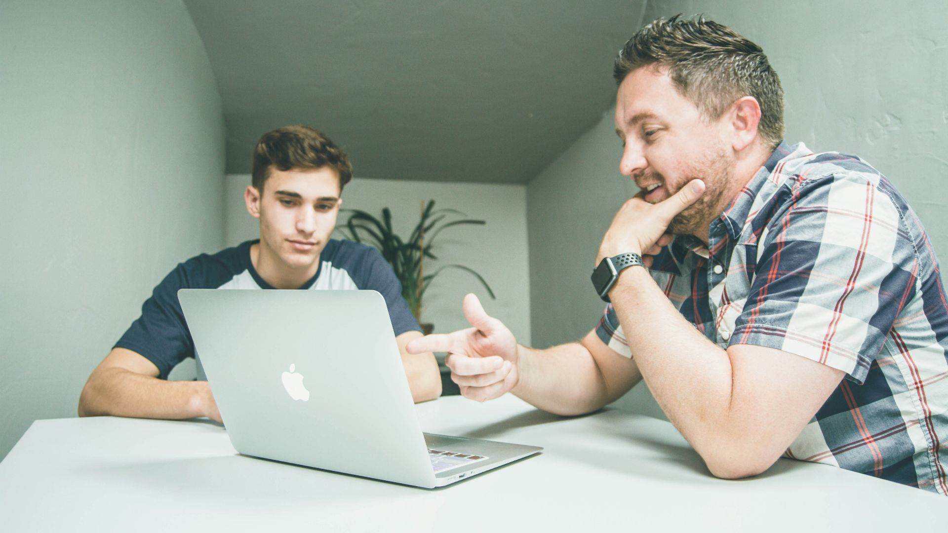 man wearing white and black plaid button-up sports shirt pointing the silver MacBook