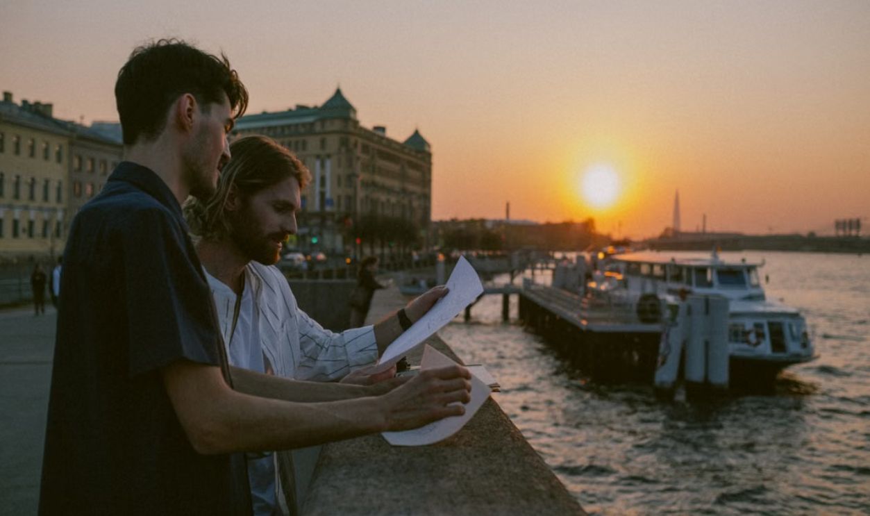 Two men reviewing papers by the riverside with a sunset backdrop, evoking an urban exploration mood.