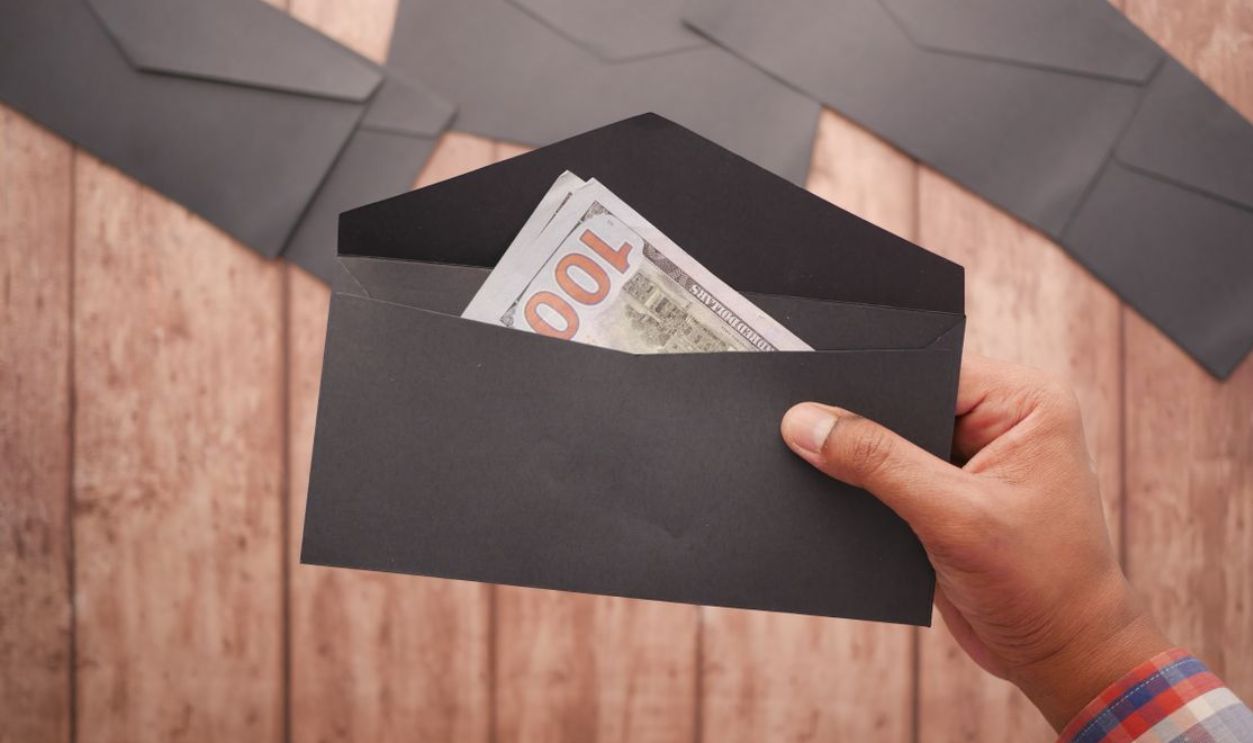 A hand holds a black envelope containing a 100 dollar bill on a wooden surface