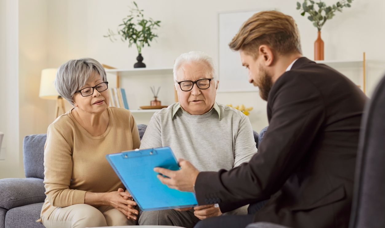 Young confident financial agent talking with senior couple about health insurance in retirement sitting on sofa at home. Elderly man and woman meeting with advisor going to sign investment contract.