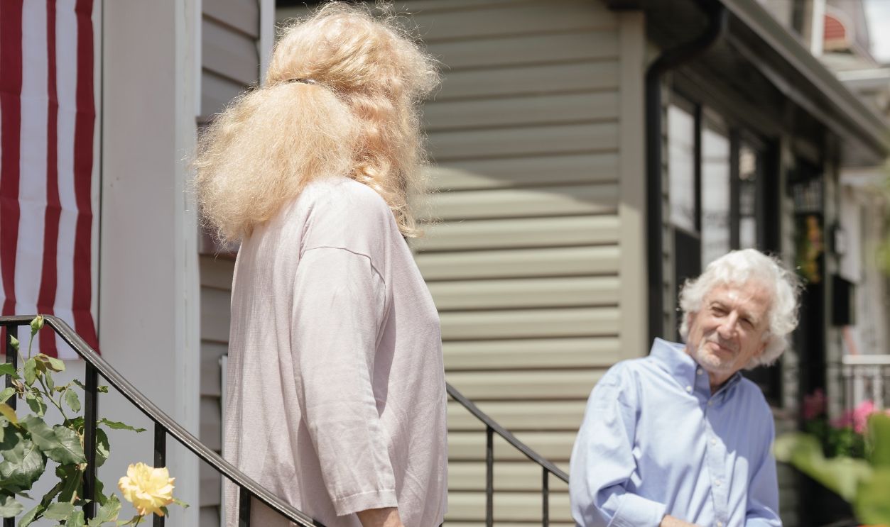 An elderly couple engaging in conversation on the porch of their suburban home with an American flag displayed