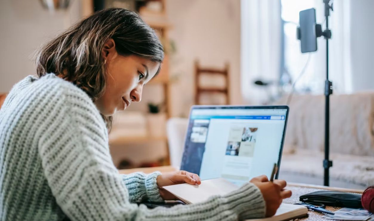 Young woman studying at home with a laptop, making notes in a cozy room during the day.