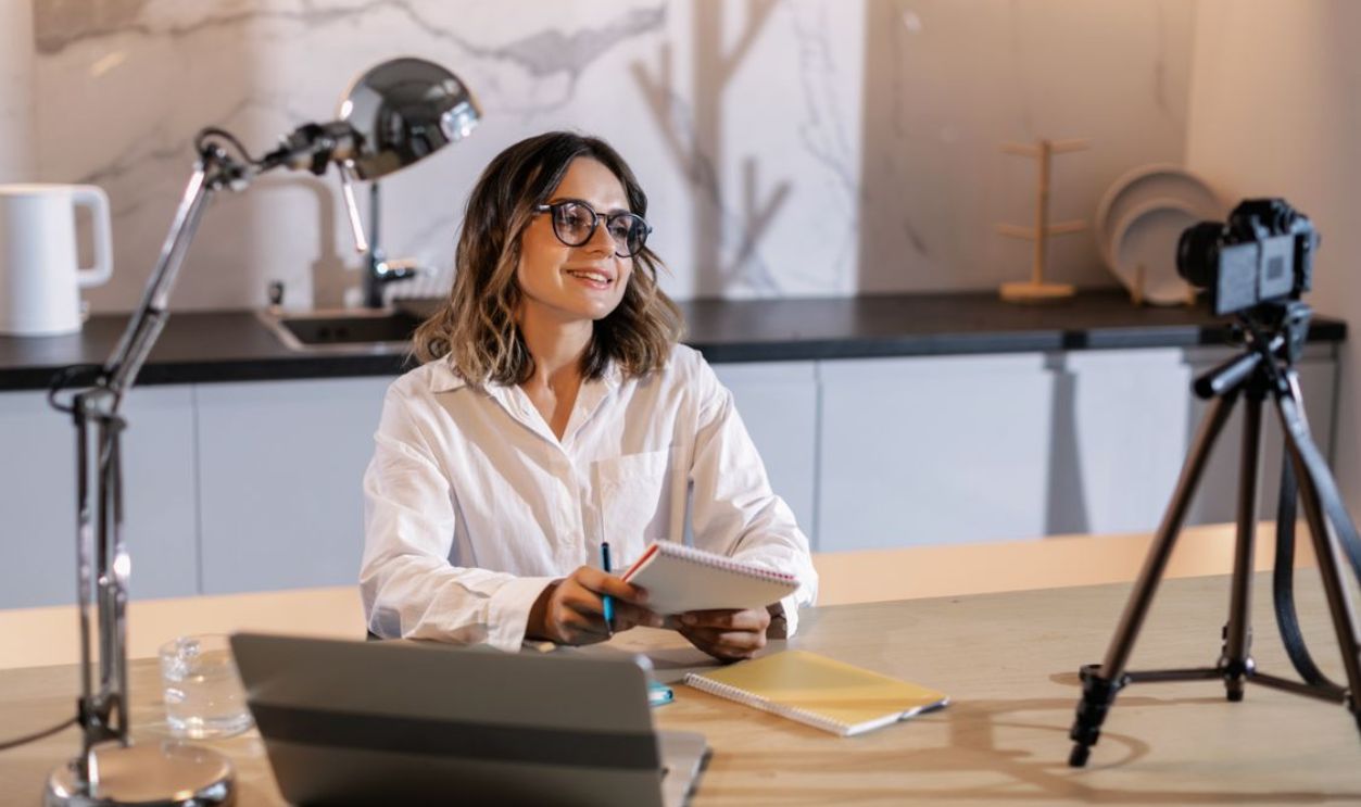 Young woman in glasses working indoors with laptop and camera, smiling while taking notes.