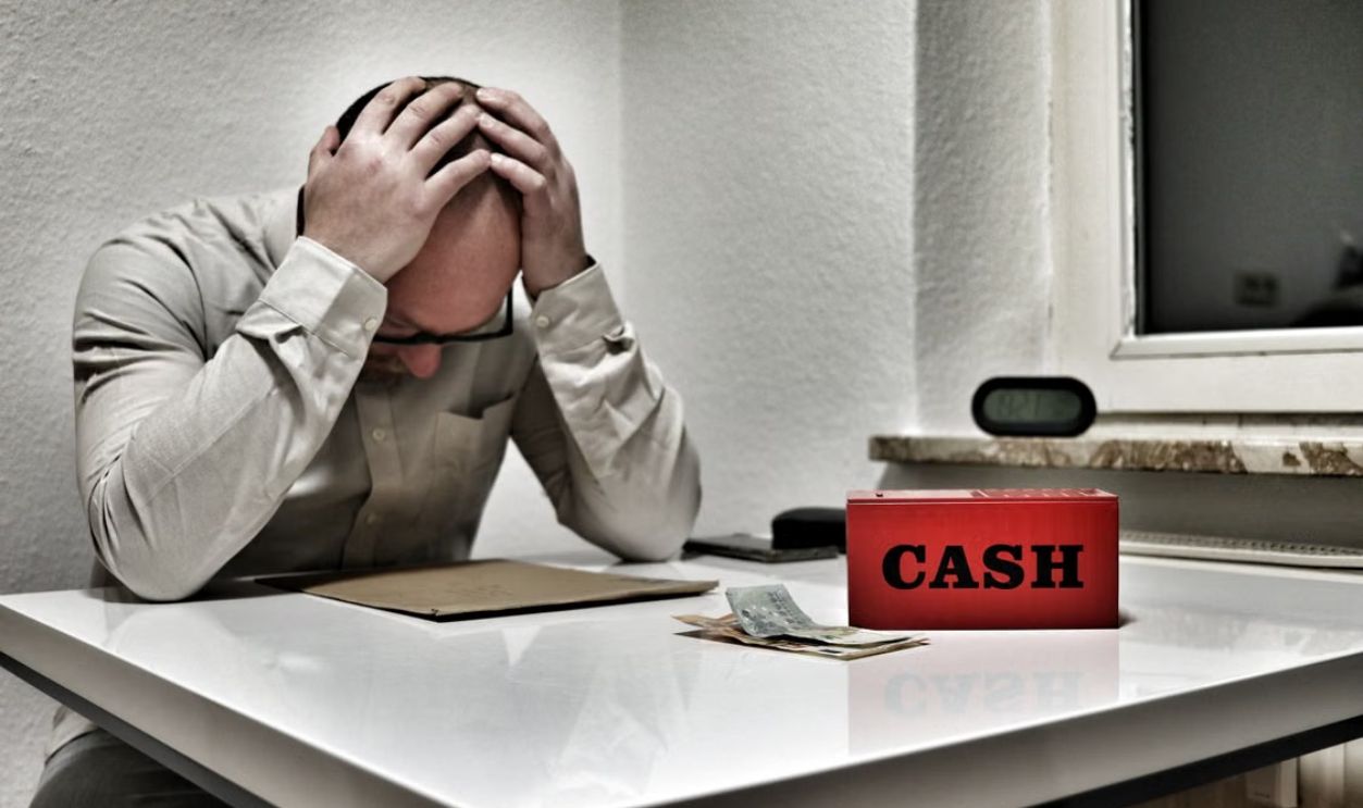 Bald man holding head in despair at desk with cashbox and money. Indoor office stress concept.