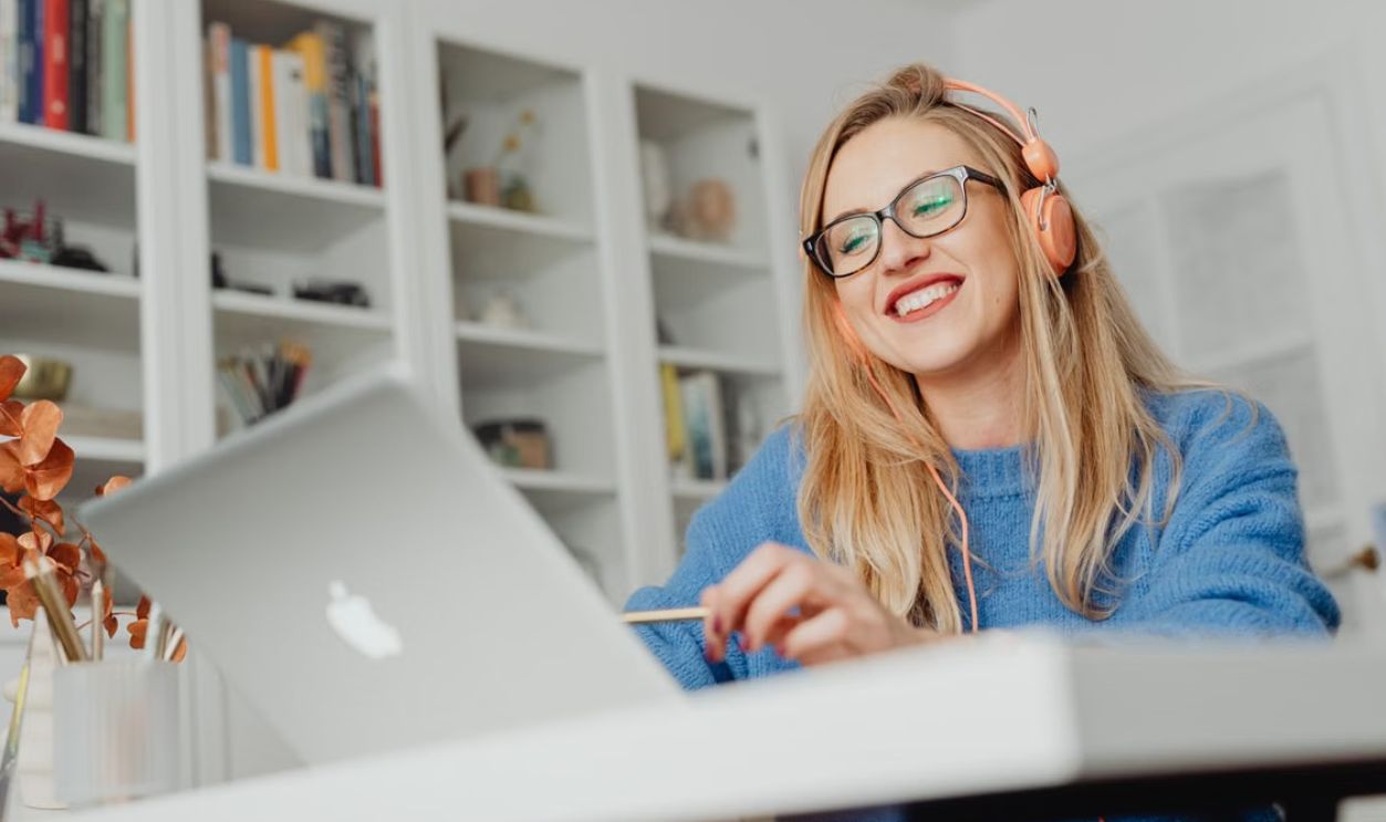 Smiling woman wearing eyeglasses and headphones, working at home on a laptop.