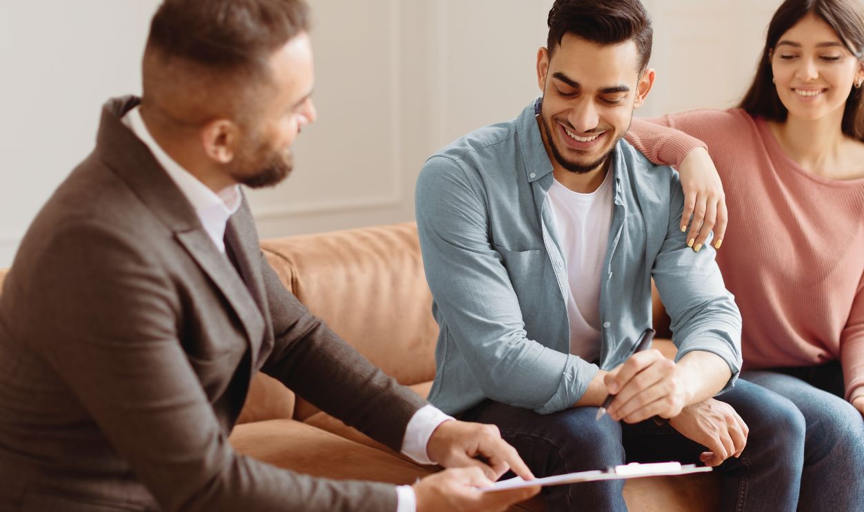 Happy Young Homeowners. Millennial Couple Buying New Private House, Financial Advisor In Suit Showing Contract, Smiling Man Signing Agreement Document, Sitting On The Couch At Office