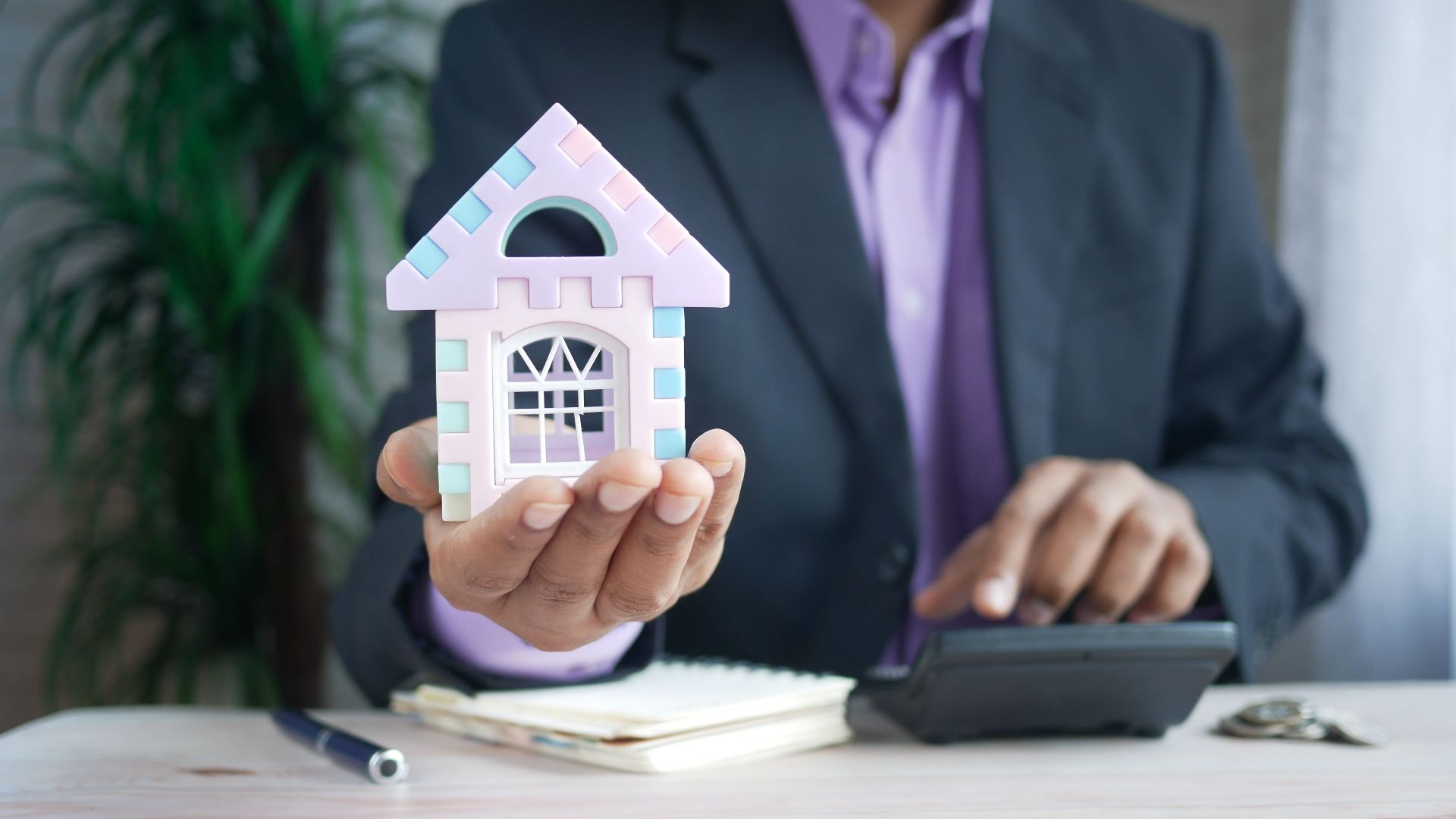 Businessman in suit holds model house, calculates real estate investments.