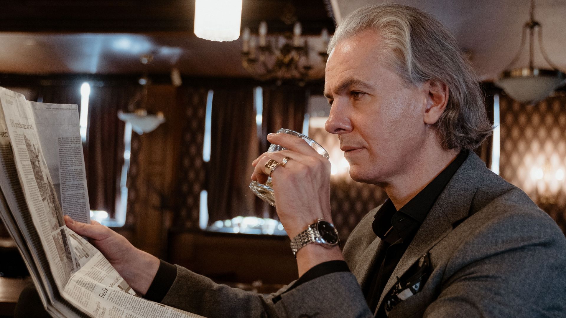 Elegant adult man in a gray suit sitting at a bar, reading a newspaper and enjoying a drink in a cozy atmosphere.